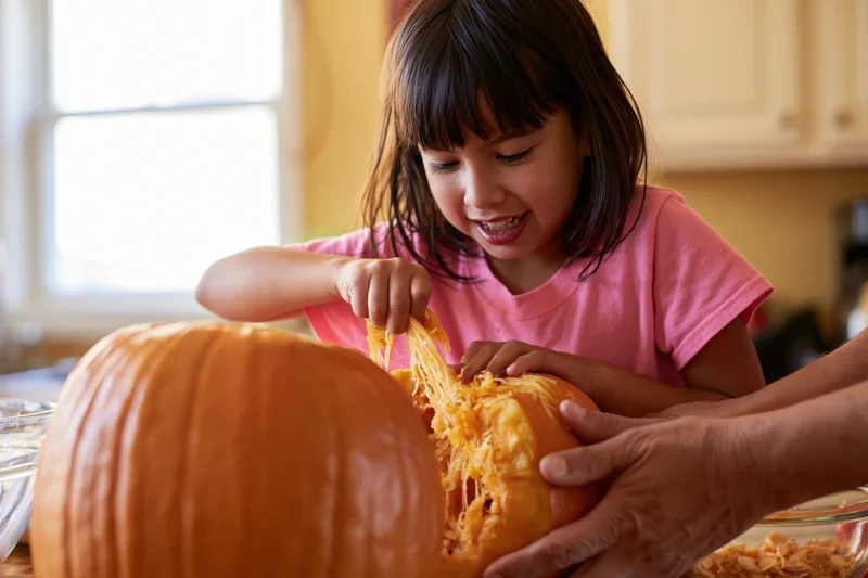 Young girl scooping stringy pumpkin guts while an adult helps in a kitchen
