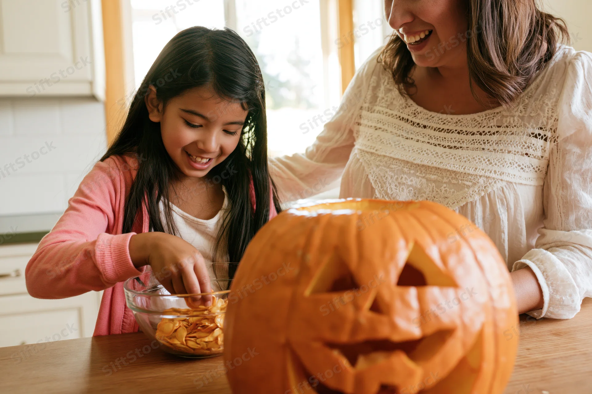 Young girl scooping pumpkin seeds into a bowl while mother smiles beside her at kitchen table with carved jack-o-lantern in foreground