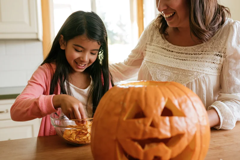 Young girl scooping pumpkin seeds into a bowl while mother smiles beside her at kitchen table with carved jack-o-lantern in foreground