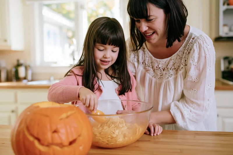 Young girl scooping pumpkin seeds into a bowl with her mother in a kitchen