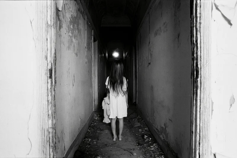 Young girl in white dress walking barefoot down a dark, decaying hallway with peeling walls and debris on the floor