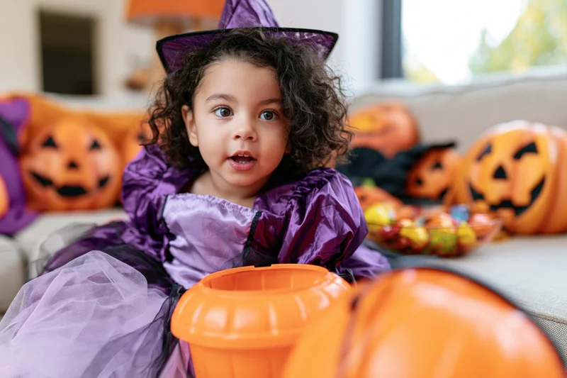 Young girl in purple witch costume sitting on couch surrounded by pumpkins and candy during halloween celebration