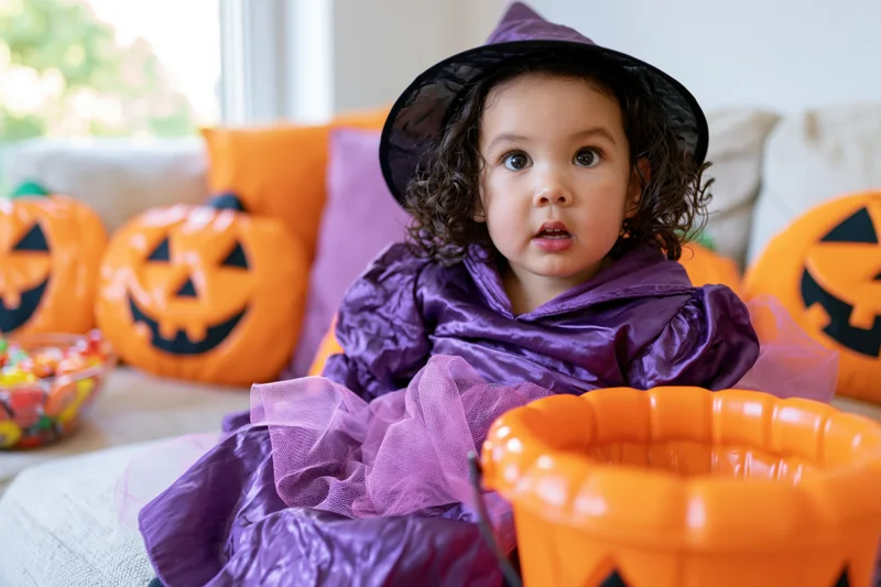 Toddler girl in purple witch costume sitting on sofa with halloween decorations and candy bucket