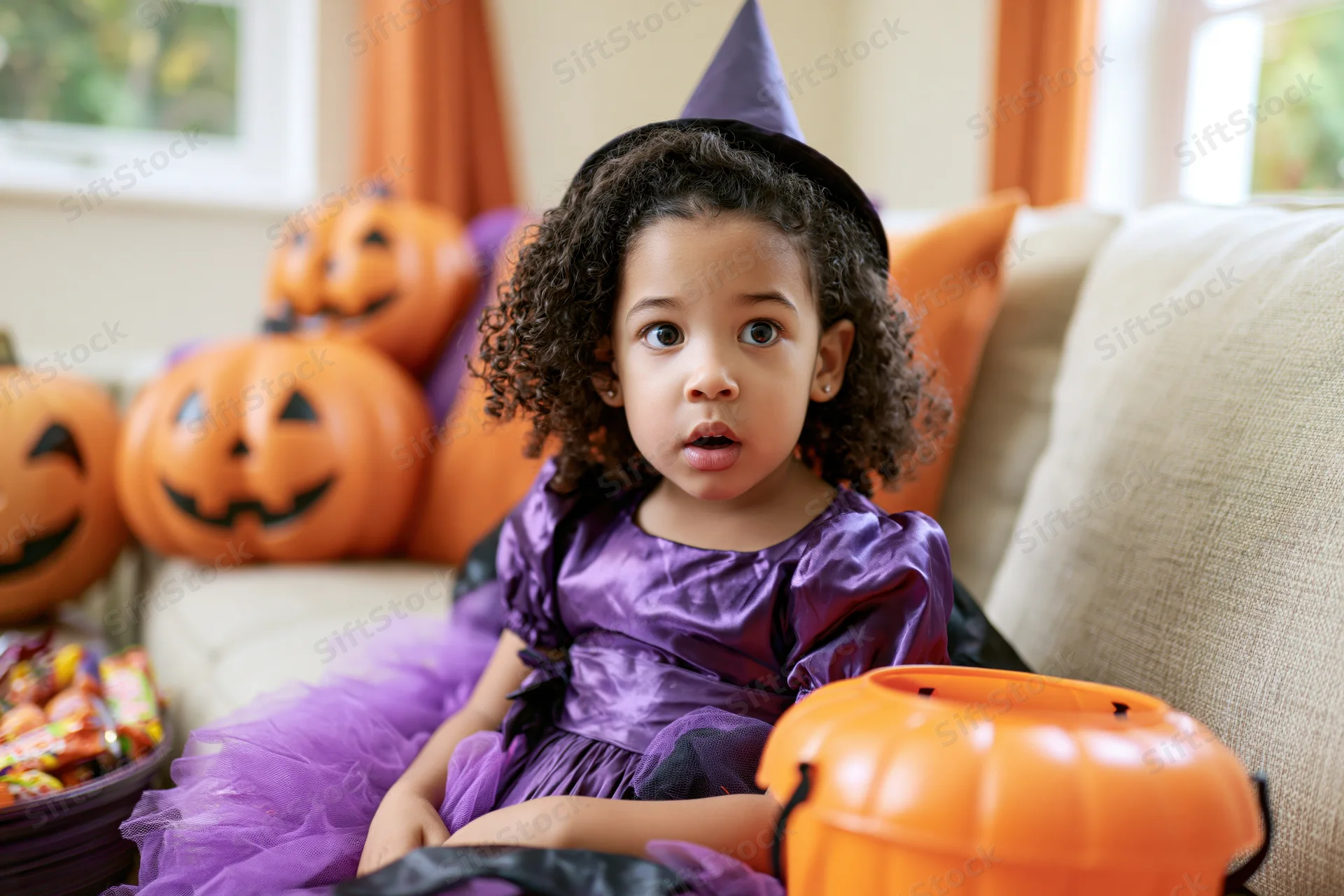Young girl in purple witch costume sitting on couch with candy bucket and pumpkins around her