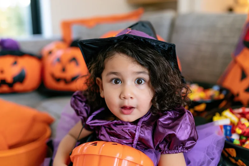 Young girl in purple witch costume holding orange bucket among halloween decorations in living room