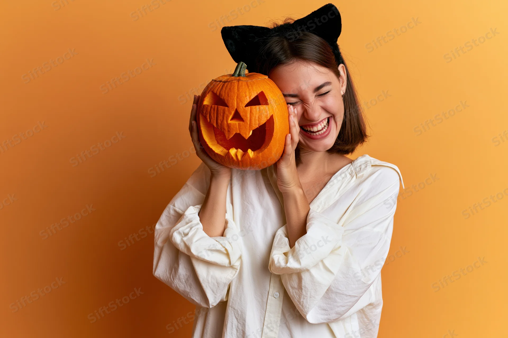 Woman laughing while holding carved jack-o-lantern to her face against orange background