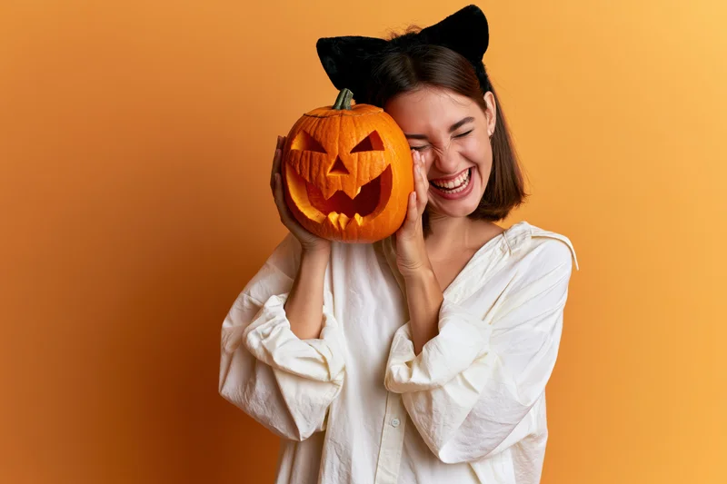 Woman laughing while holding carved jack-o-lantern to her face against orange background