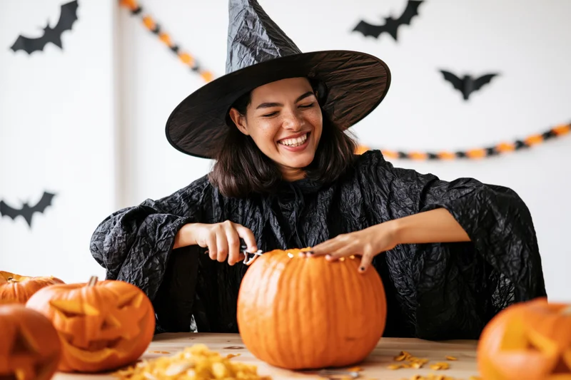 Woman in witch costume carving pumpkin at table with halloween decorations