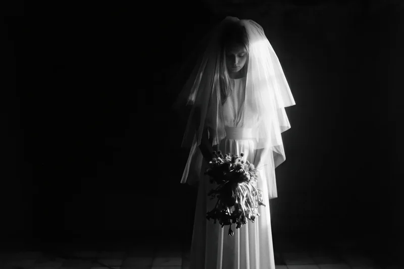 Bride in white wedding dress and long veil holding bouquet, standing in dark studio with dramatic lighting