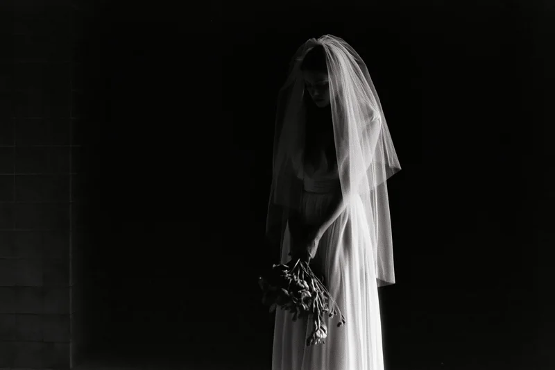 Bride in white wedding dress and long veil holding dried bouquet, standing in dark studio with dramatic lighting