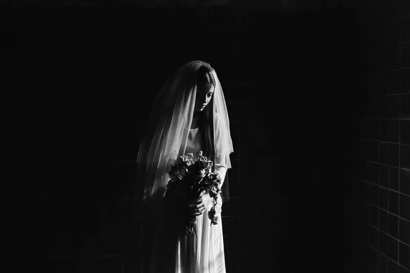 Bride in white wedding dress and veil holding bouquet, standing against dark tiled wall