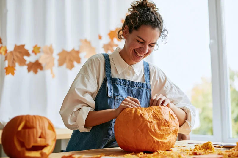 Woman carving a pumpkin at a kitchen table with autumn decorations in the background