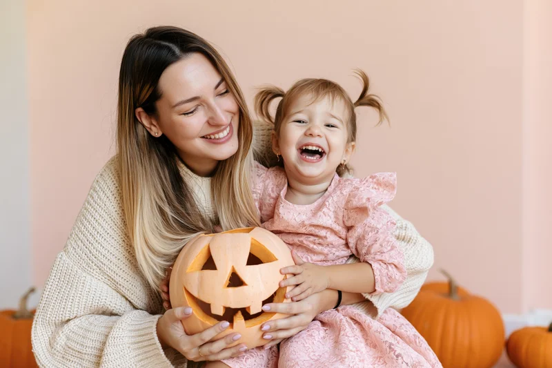 Woman hugging laughing toddler holding carved pumpkin indoors