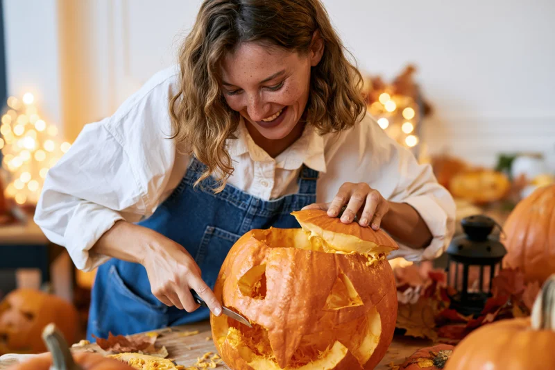 Woman smiling while carving a pumpkin on a kitchen table surrounded by autumn decorations