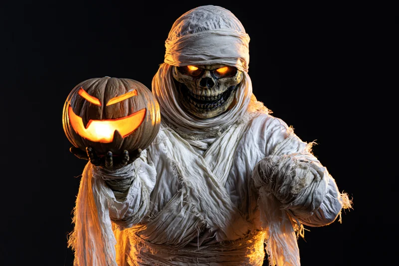 Mummy in wrapped costume holding glowing jack-o-lantern against black background