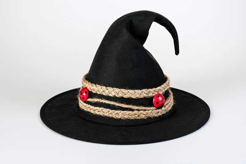 Black fabric witch hat with curved point and twine band decorated with red ladybug buttons on white background