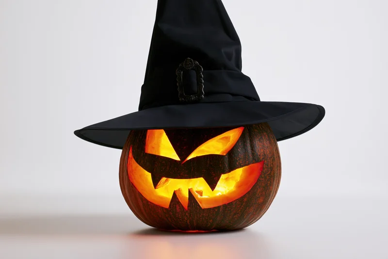 Carved jack-o-lantern with glowing face wearing a tall black witch hat on a white surface