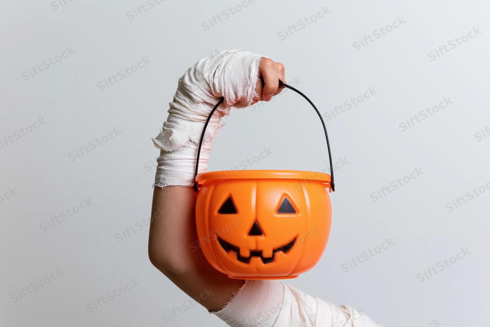 Child's arm wrapped in tattered white mummy bandages holding an orange plastic jack-o-lantern bucket for trick-or-treating