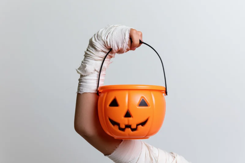 Child's arm wrapped in tattered white mummy bandages holding an orange plastic jack-o-lantern bucket for trick-or-treating