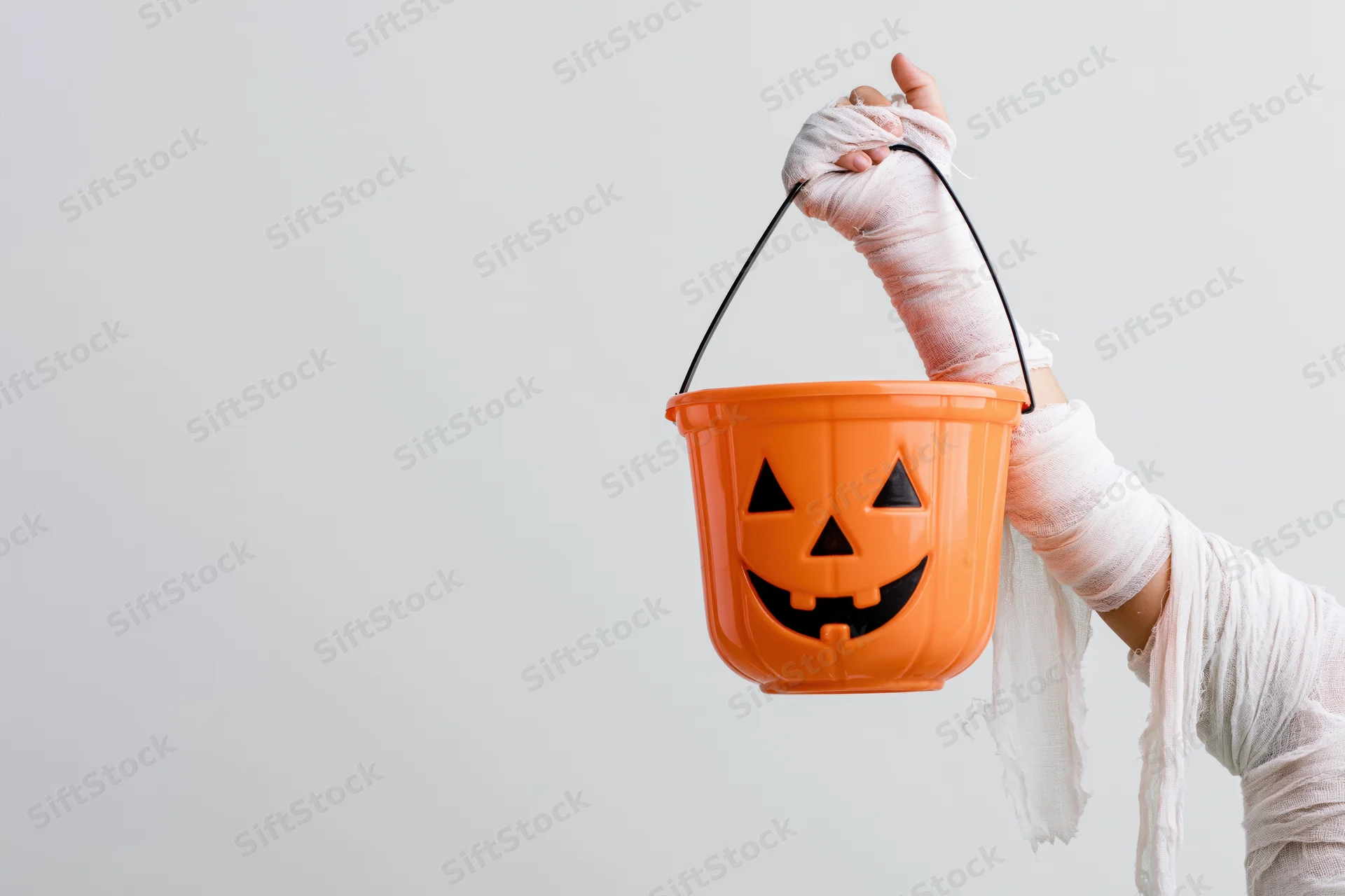 Child's arm wrapped in white mummy bandages holding an orange jack-o-lantern bucket against a plain background
