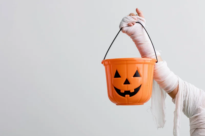 Child's arm wrapped in white mummy bandages holding an orange jack-o-lantern bucket against a plain background