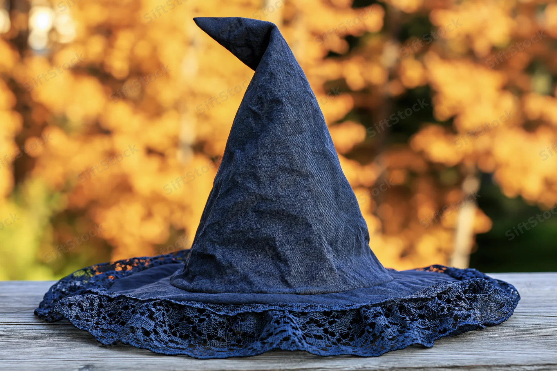 Dark navy fabric witch hat with pointed cone and lace trim resting on wooden table outdoors in autumn foliage