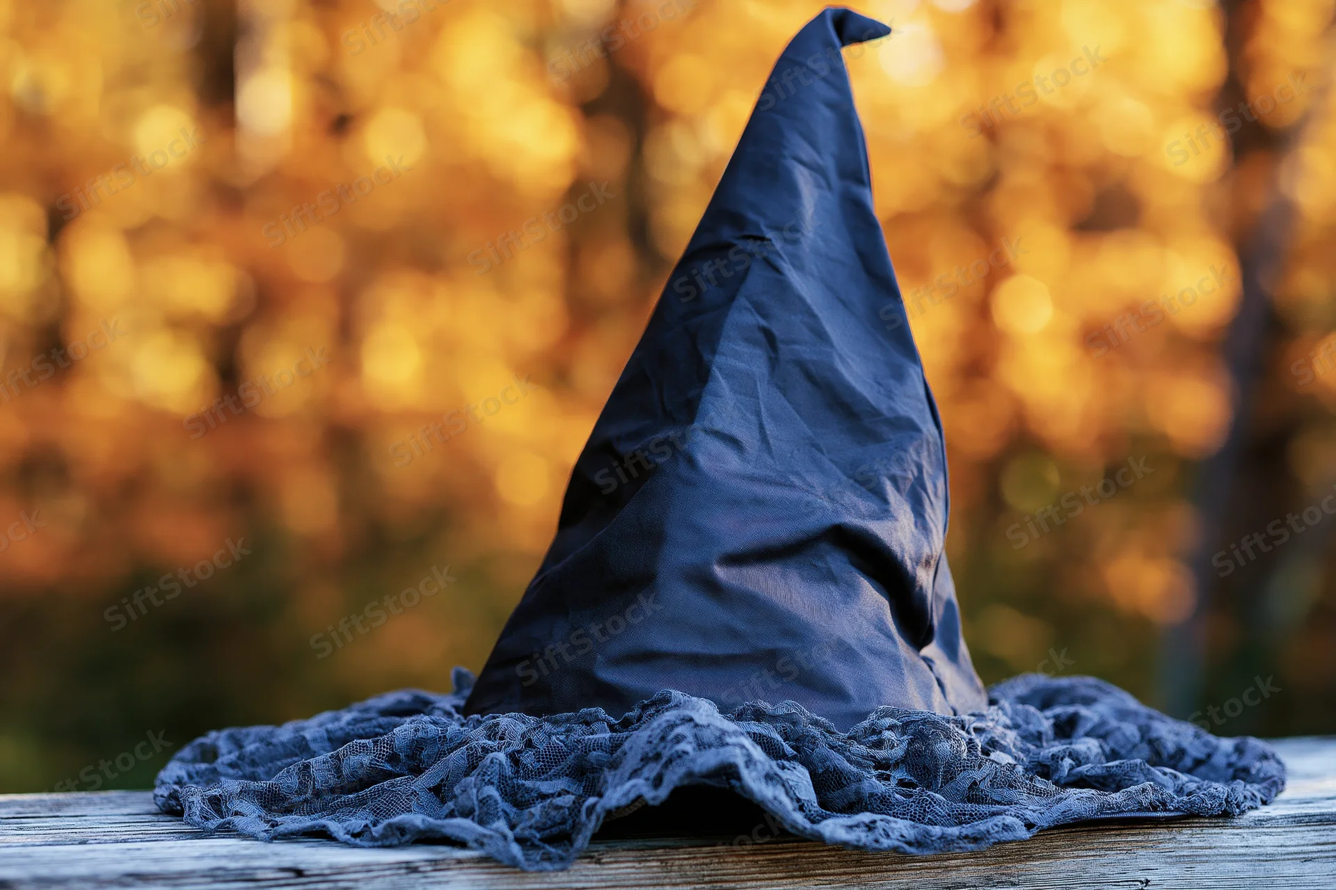 Dark navy witch hat with ruffled lace brim resting on wooden surface against golden autumn foliage background