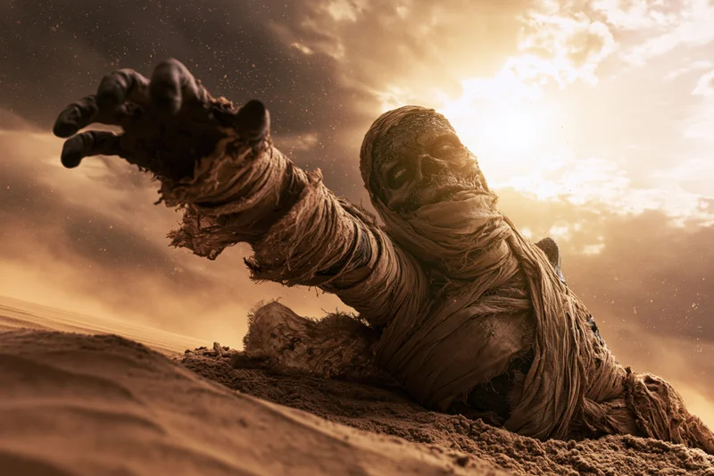 Decayed mummy emerging from desert sand with arm raised toward sunlit sky