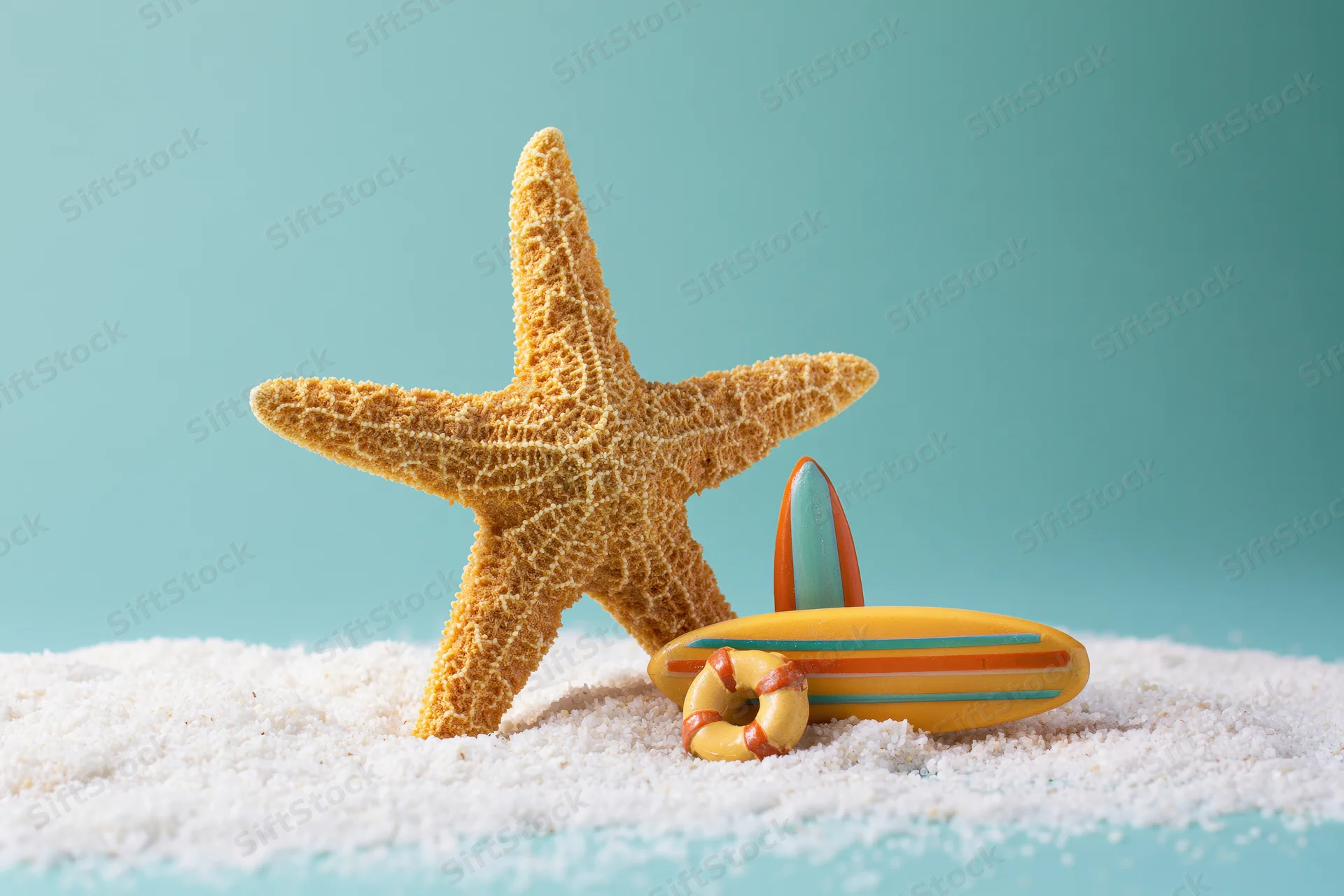Dried starfish standing upright in white sand beside a toy surfboard and lifebuoy on a teal background