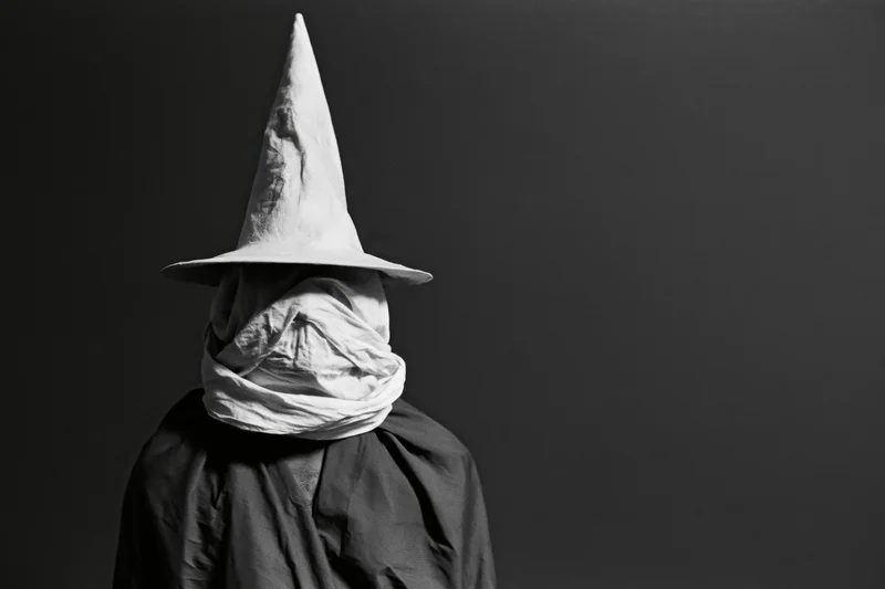 Hooded figure in pointed witch hat with face covered by cloth, standing against dark background