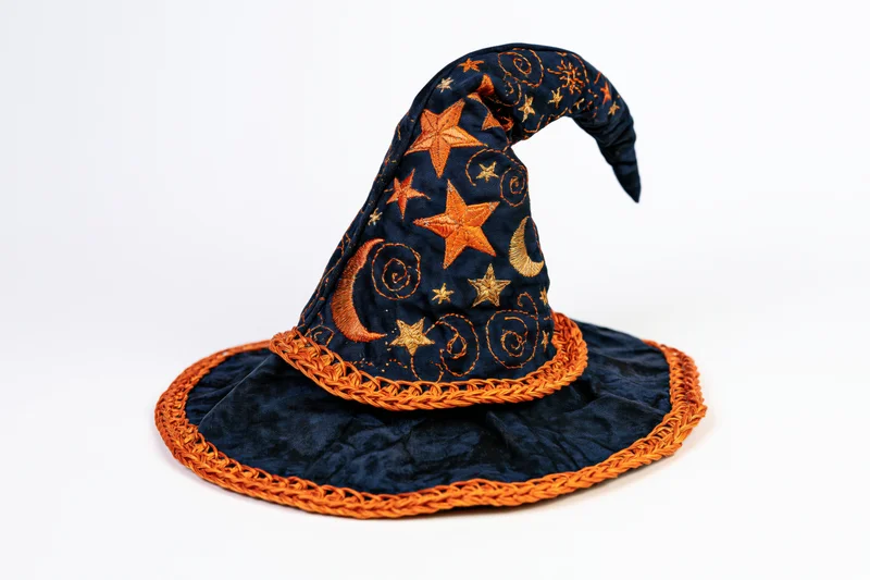 Handmade fabric witch hat with embroidered stars and moons, curved tip, orange braided trim, lying on white background