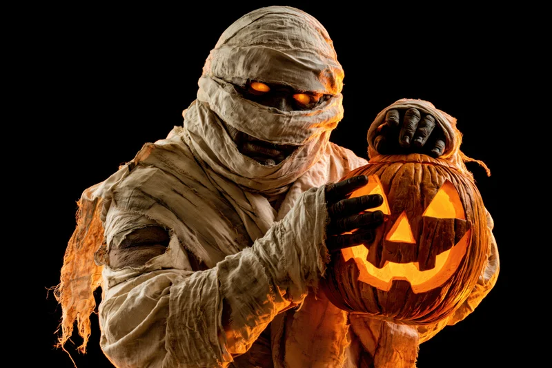 Mummy in linen bandages holding a glowing carved pumpkin against black background