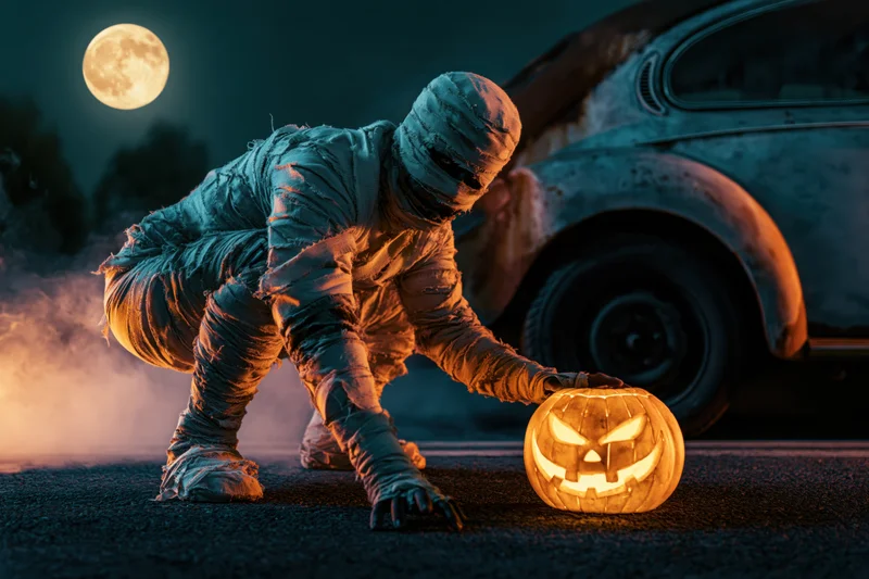 Mummy in costume crouching and reaching toward a glowing jack-o-lantern on asphalt at night near a rusted vintage car under a full moon