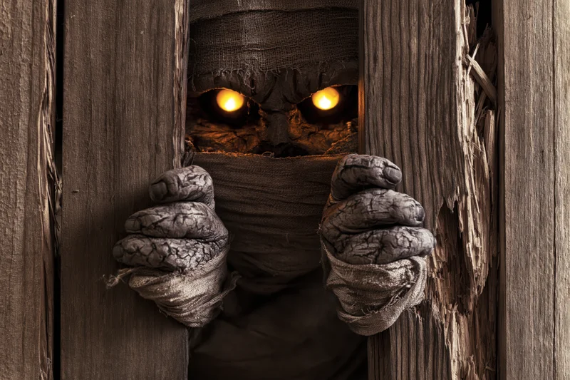 Mummy creature with glowing amber eyes peering through a cracked wooden door