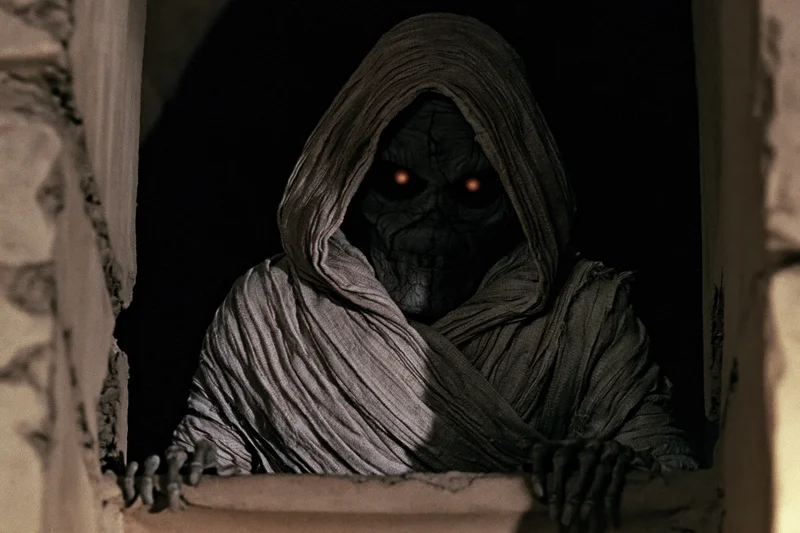 Mummy creature with glowing red eyes leaning forward through a broken window frame in a dark ruin