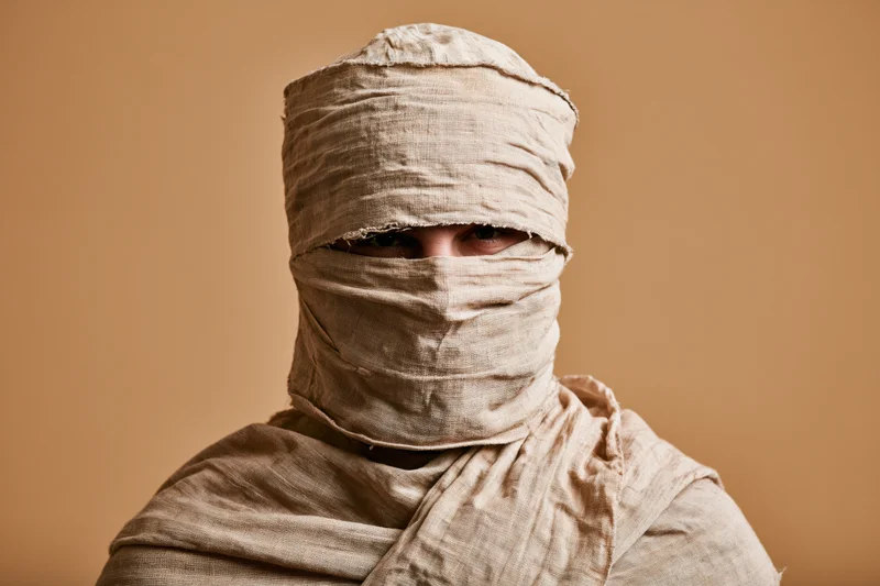 Person wrapped in aged linen cloth from head to shoulders, eyes visible, in studio setting