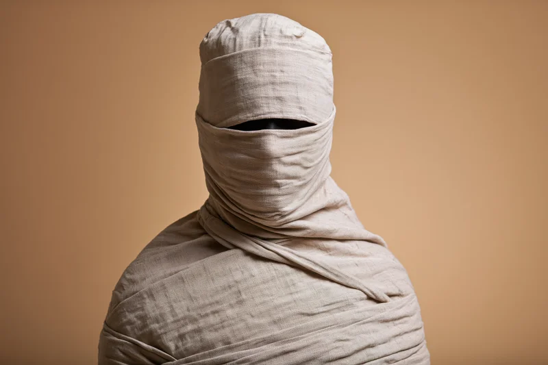 Person wrapped in linen bandages from head to shoulders against a plain beige background