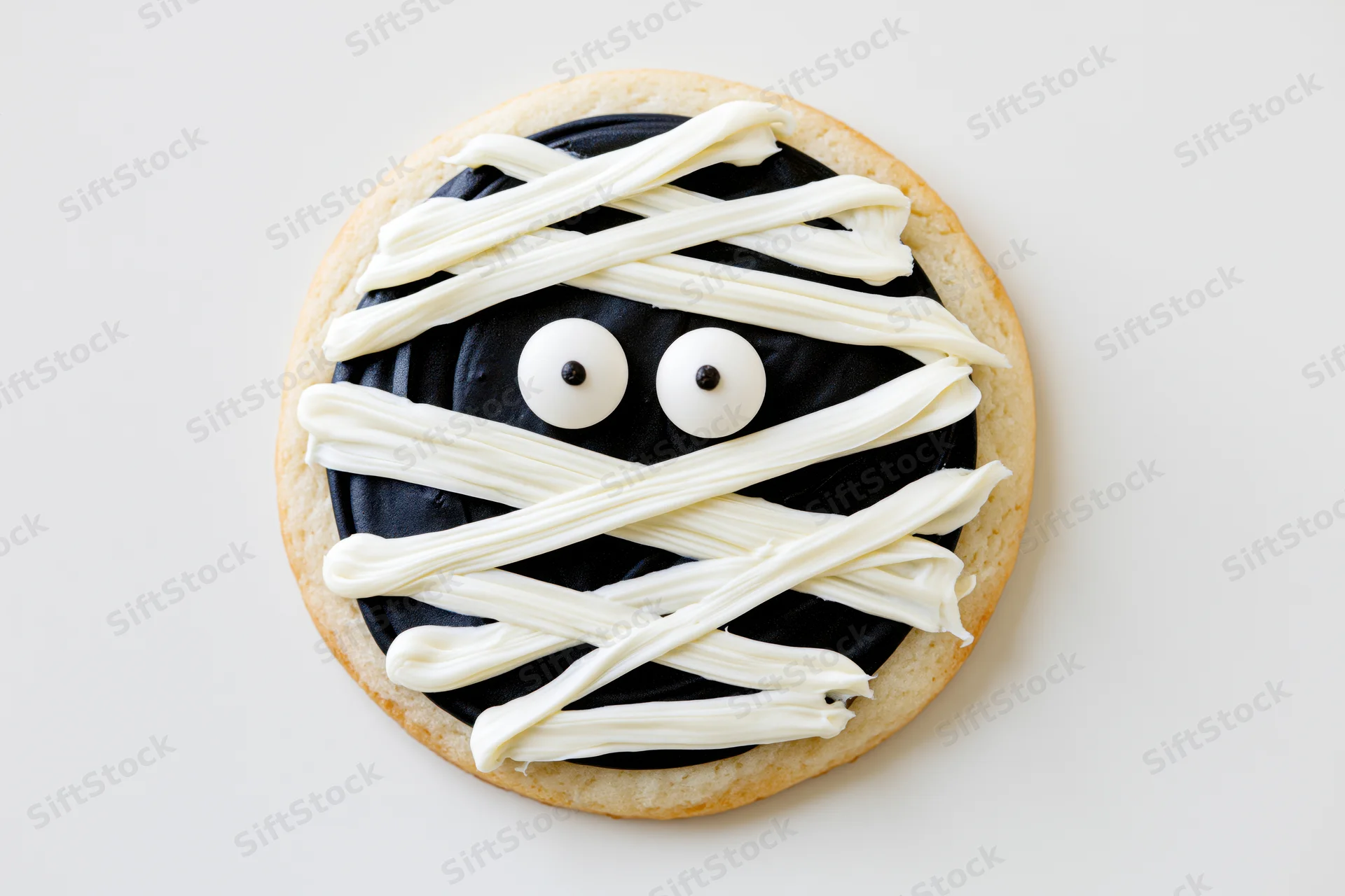 Round sugar cookie decorated as a mummy face with white frosting and candy eyes on white background
