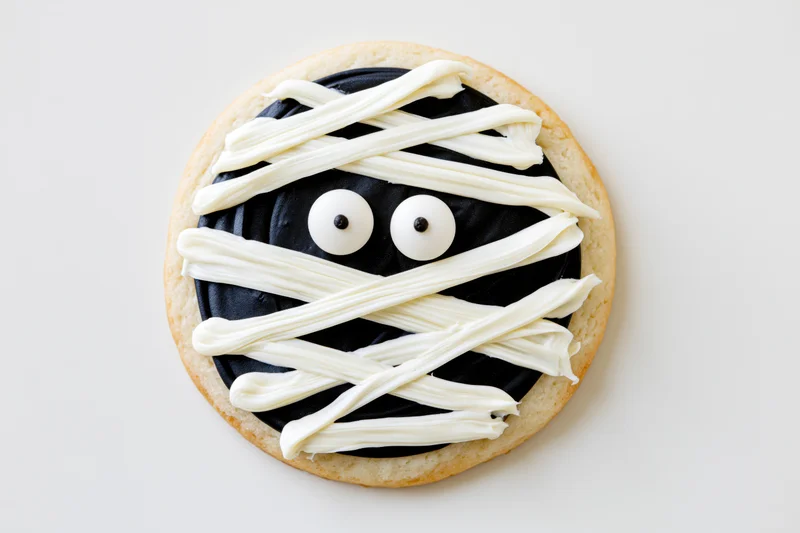 Round sugar cookie decorated as a mummy face with white frosting and candy eyes on white background