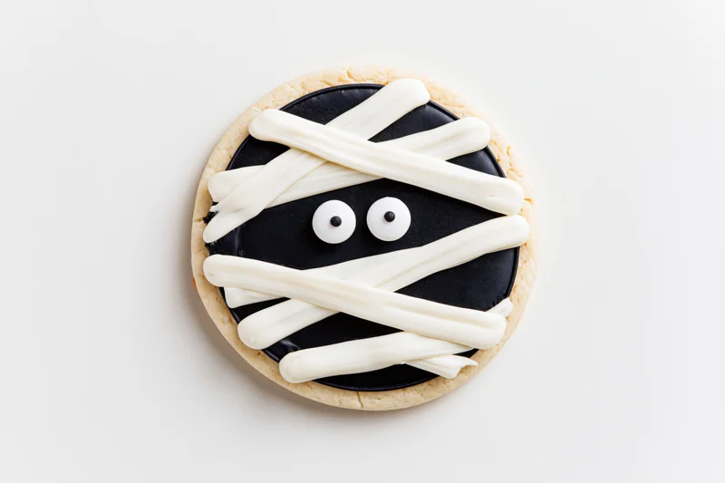 Round sugar cookie decorated as a mummy face with white frosting bandages and candy eyes on white background