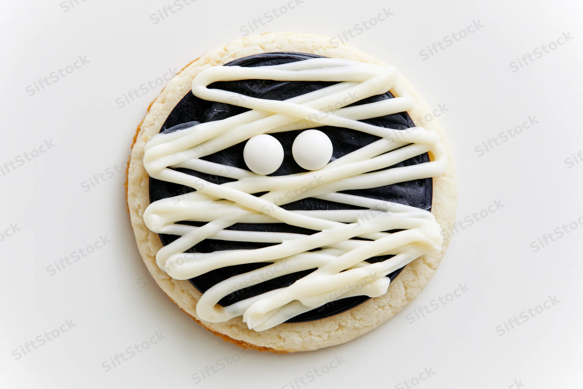 Round sugar cookie decorated as a mummy face with white frosting and candy eyes on white background