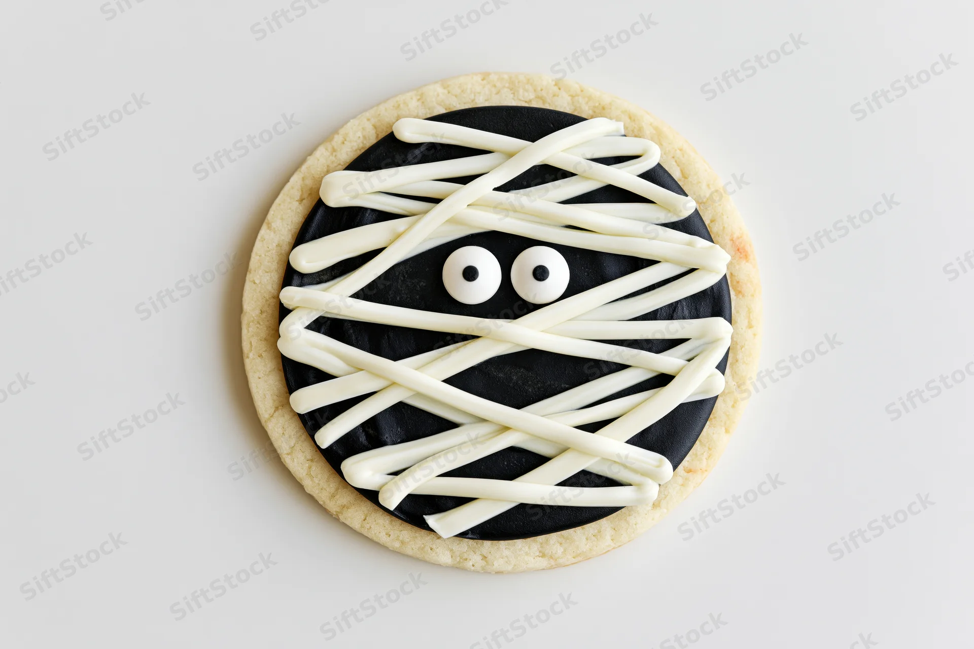 Round sugar cookie decorated as a mummy face with white frosting and candy eyes on white surface