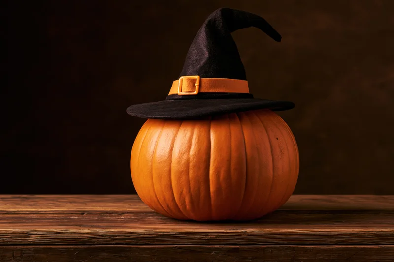Round orange pumpkin wearing black witch hat on worn wooden table