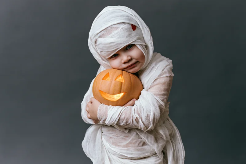 Child in mummy costume hugging a glowing jack-o-lantern against dark gray background