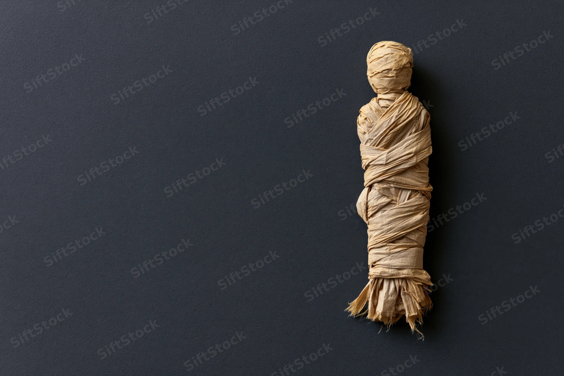 Handmade mummy figure wrapped in linen strips on dark surface with copyspace