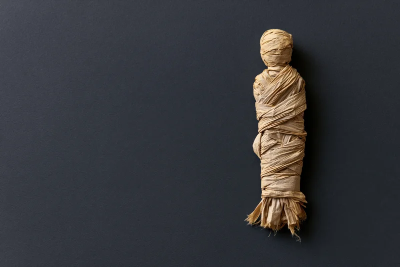 Handmade mummy figure wrapped in linen strips on dark surface with copyspace