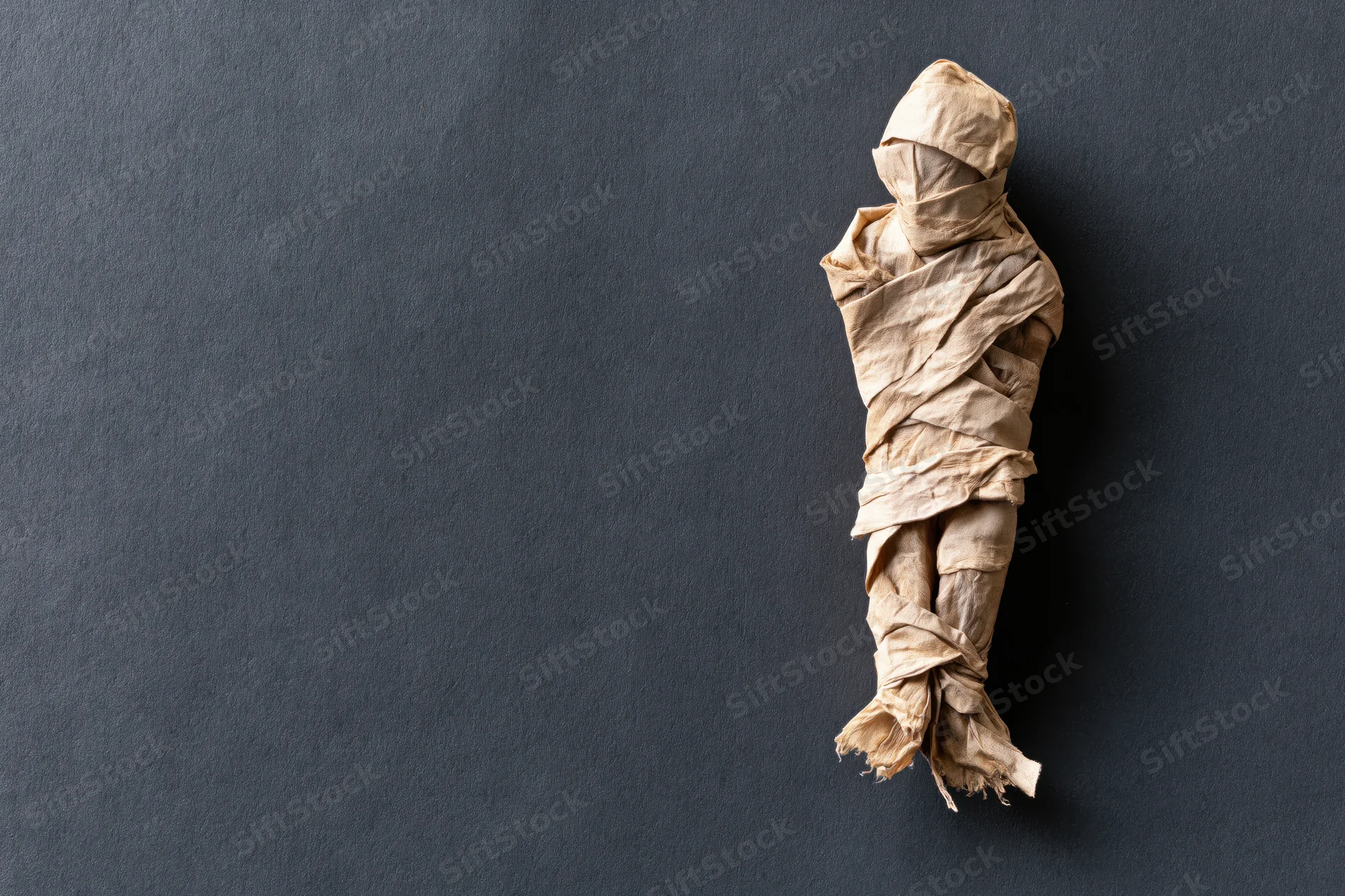 Handmade mummy figure wrapped in paper strips on dark textured surface with copyspace
