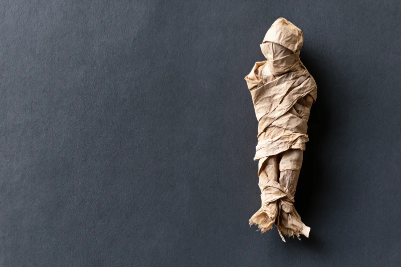 Handmade mummy figure wrapped in paper strips on dark textured surface with copyspace