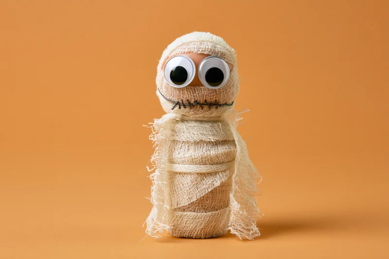 Handmade mummy figurine wrapped in cream gauze with googly eyes on orange background