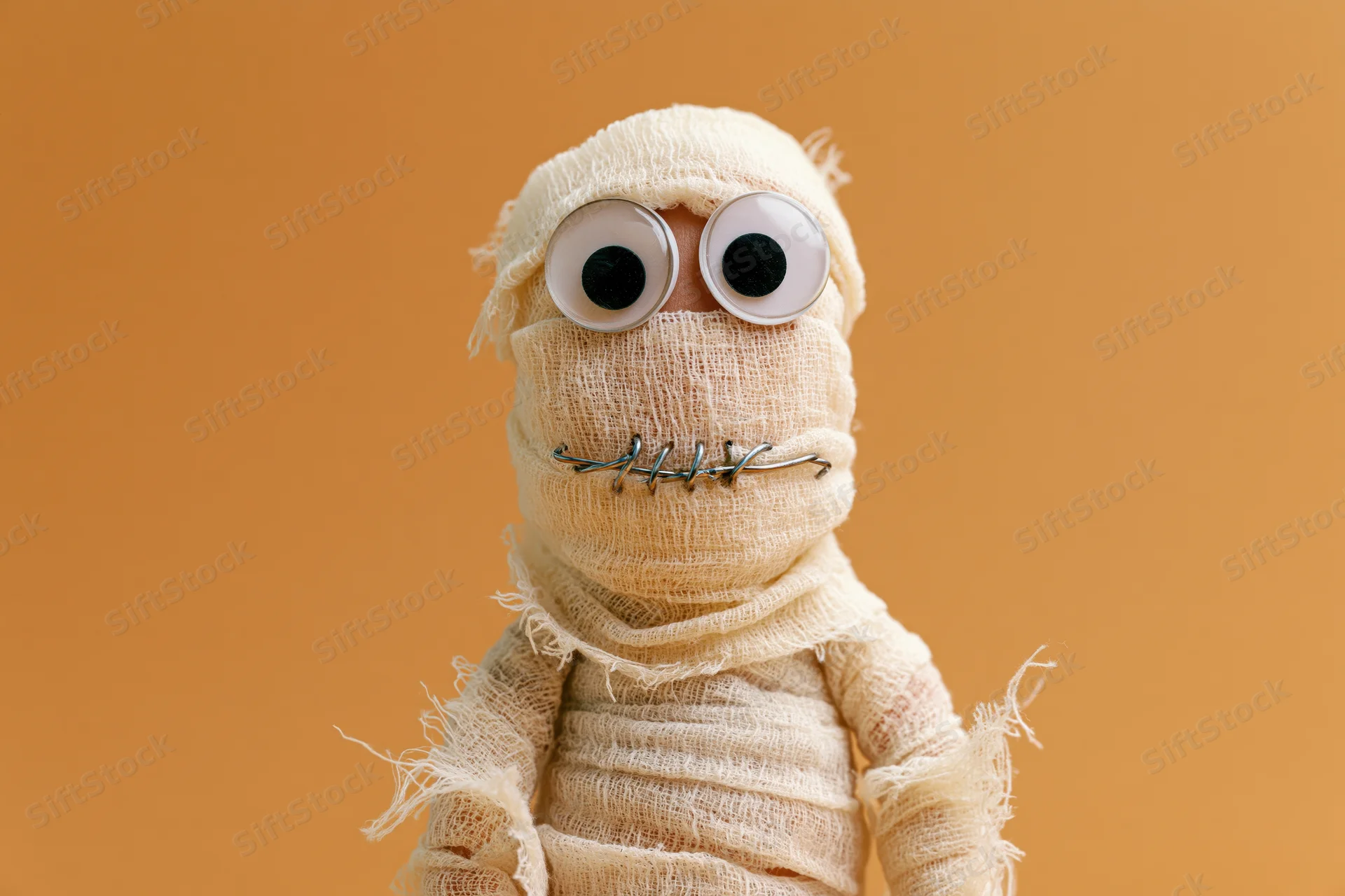 Handmade mummy figurine wrapped in cream gauze with googly eyes and wire mouth against orange background
